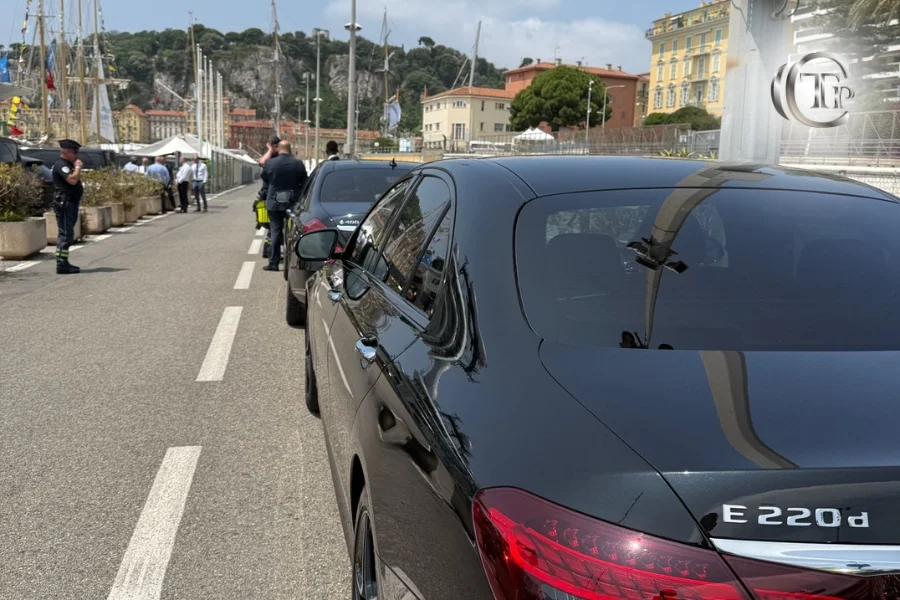 Rear shot of black Mercedes E-Class with convoy in the background and police presence at the port of Nice.