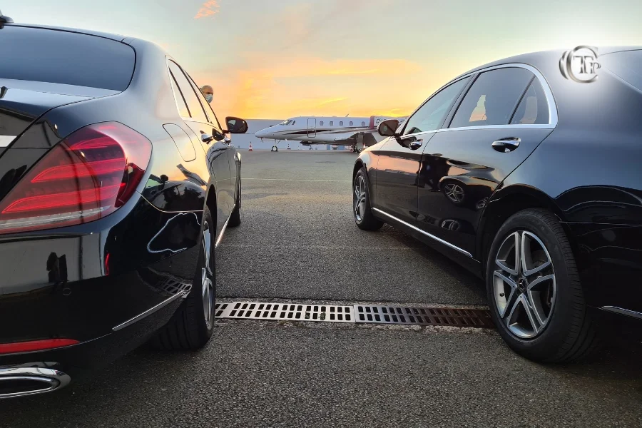 Deux Mercedes noires garées sur le tarmac de l’aéroport avec un jet privé en arrière-plan au coucher du soleil.